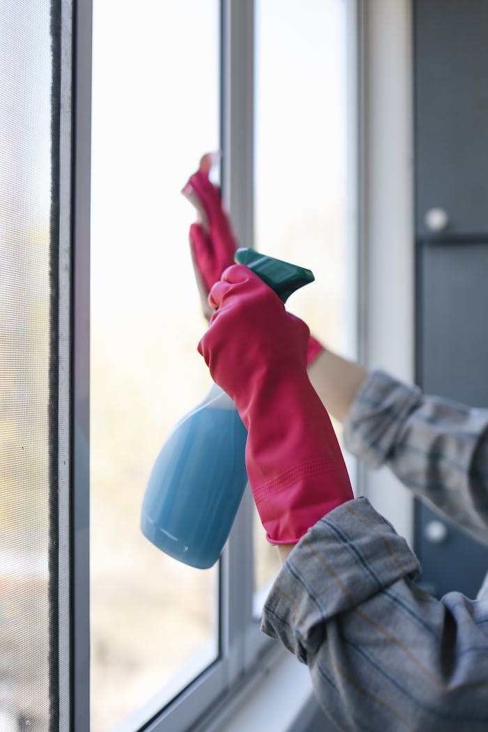 hero-02 Hands in rubber gloves using a spray bottle to clean a window indoors.